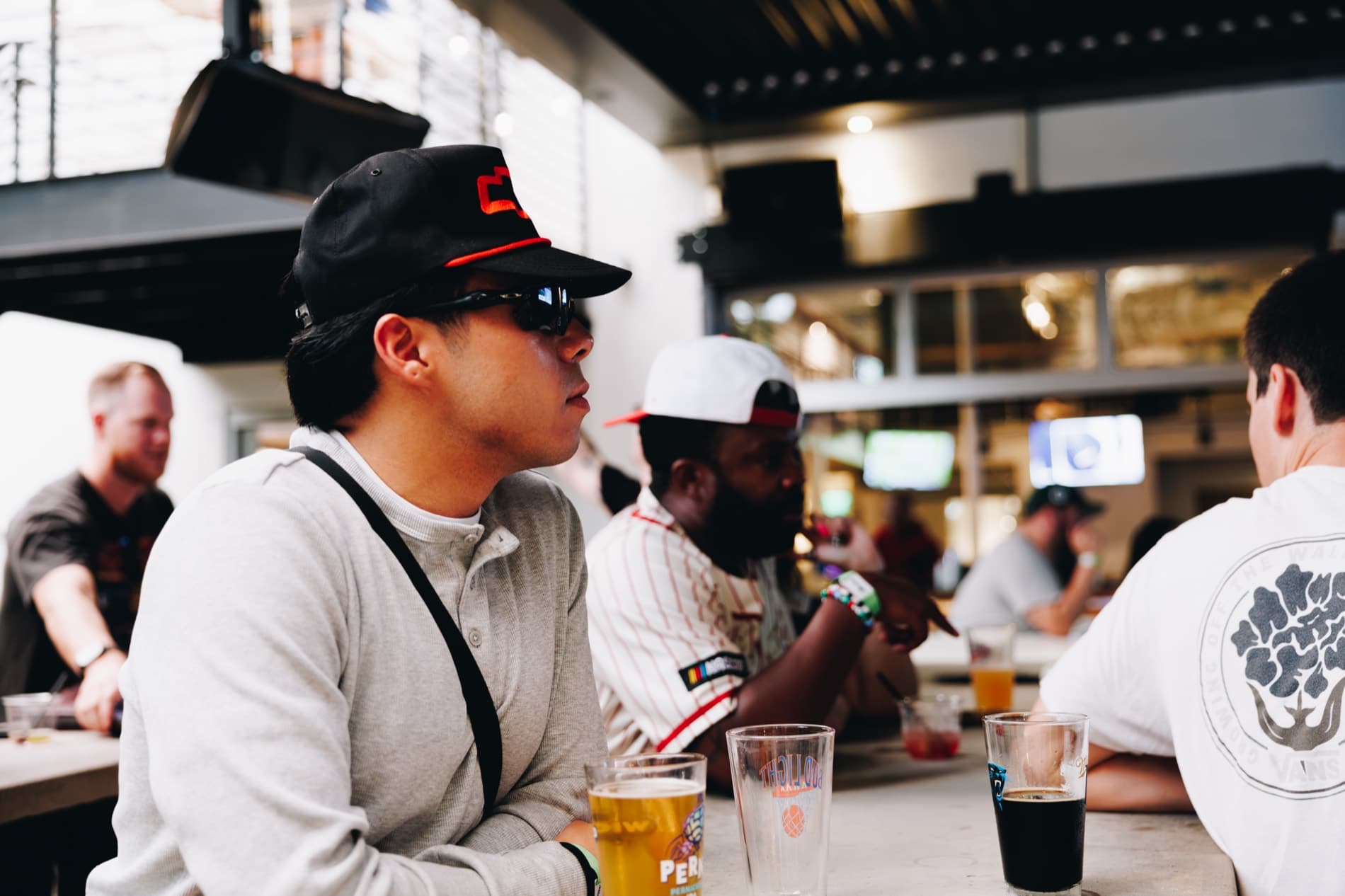 A man in a black cap and sunglasses sits at a bar with other patrons and drinks on the counter.