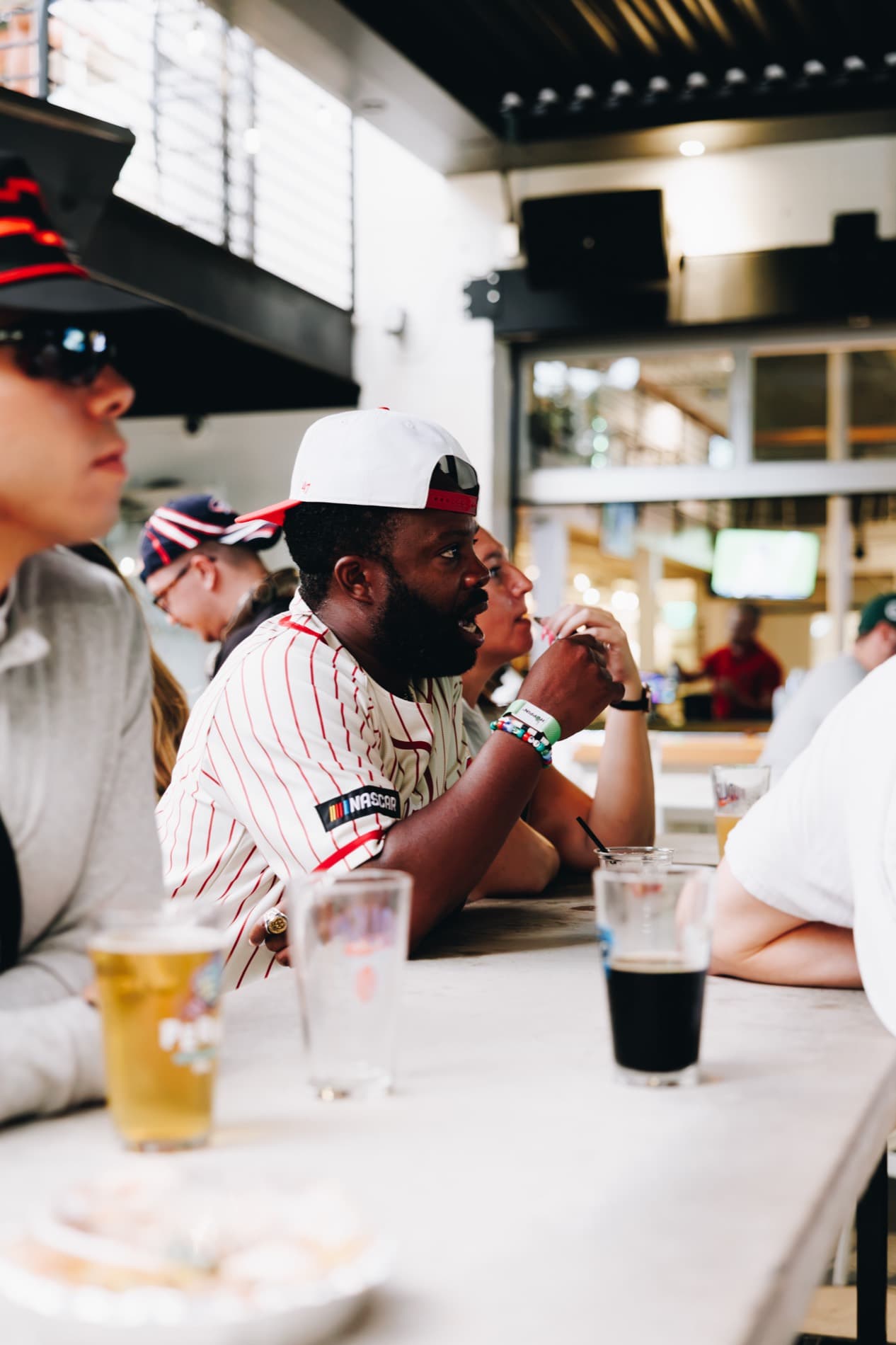 Fans sit at a bar table with drinks, including a man in a pinstriped shirt with a NASCAR patch and a backward white cap.
