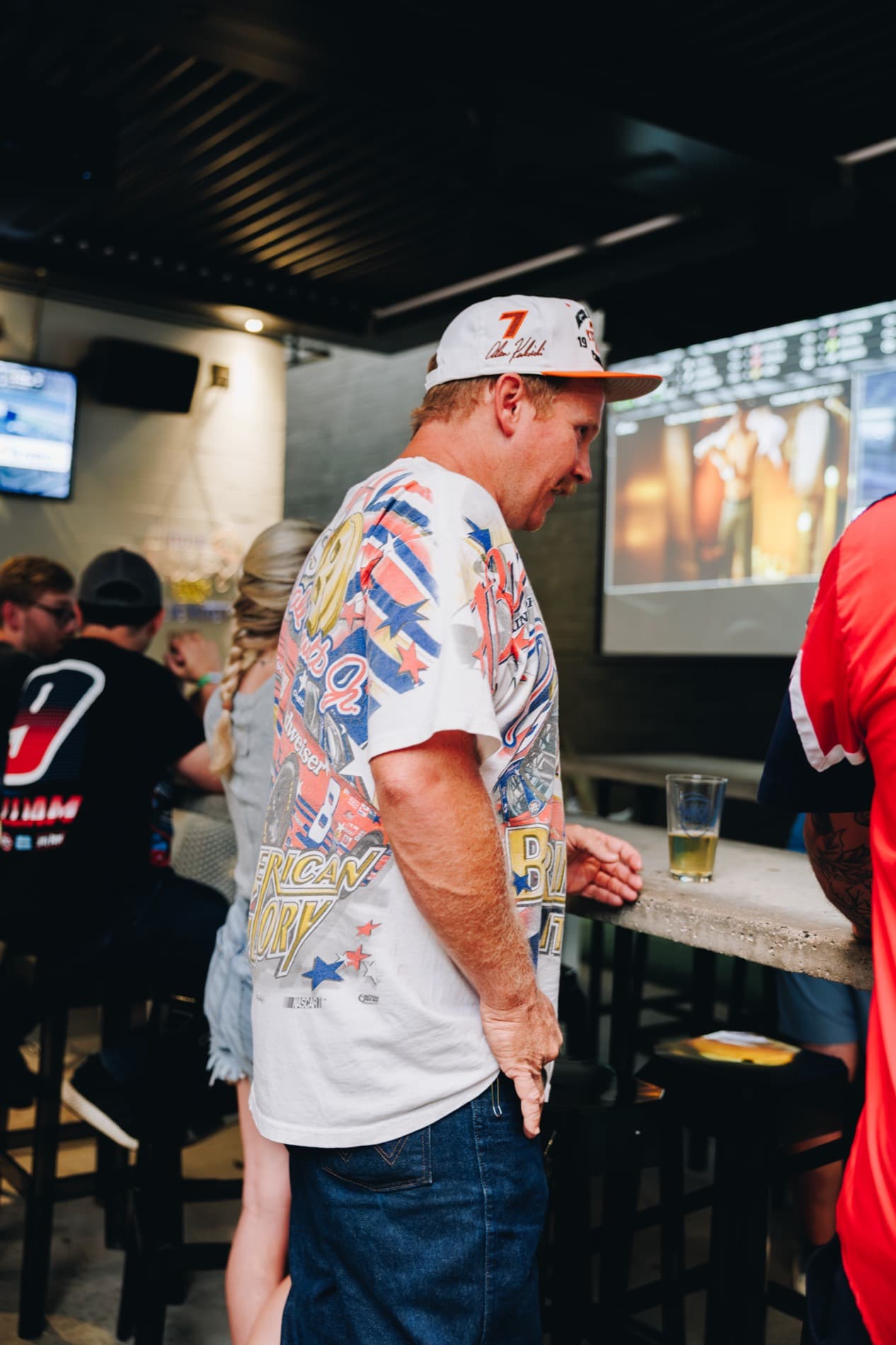 A man wearing a white NASCAR racing T-shirt and a white number 7 cap stands at a bar with a drink while people sit nearby and a screen plays in the background.