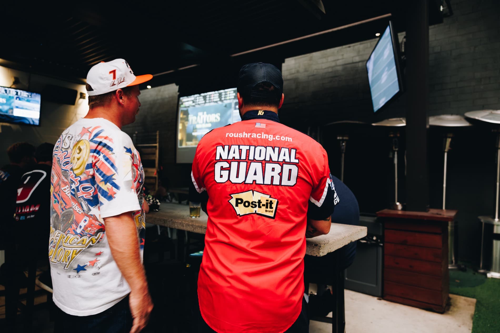 Two NASCAR fans stand in a bar area watching wall-mounted screens, including one wearing a red National Guard Roush Racing shirt.