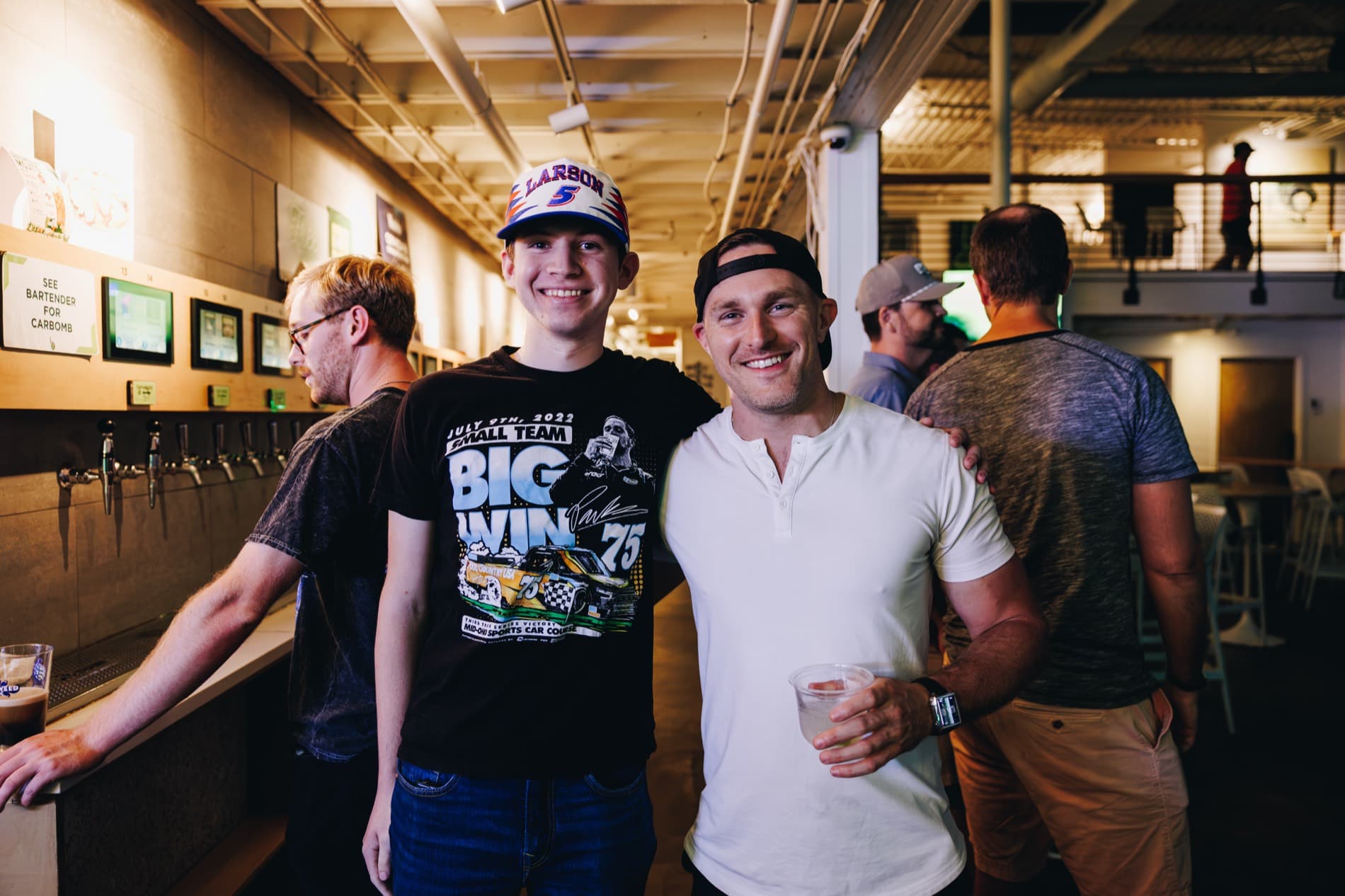 Two men smile with their arms around each other inside a bar, with beer taps and other patrons in the background.