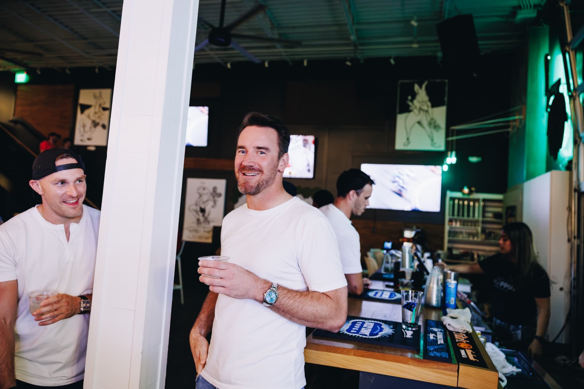 Two men in white shirts hold drinks and smile inside a bar with televisions and staff behind the counter.