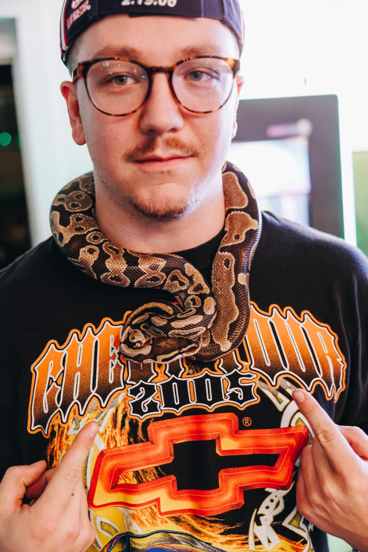 A man wearing glasses and a racing-themed Chevrolet shirt has a patterned snake draped around his neck indoors.