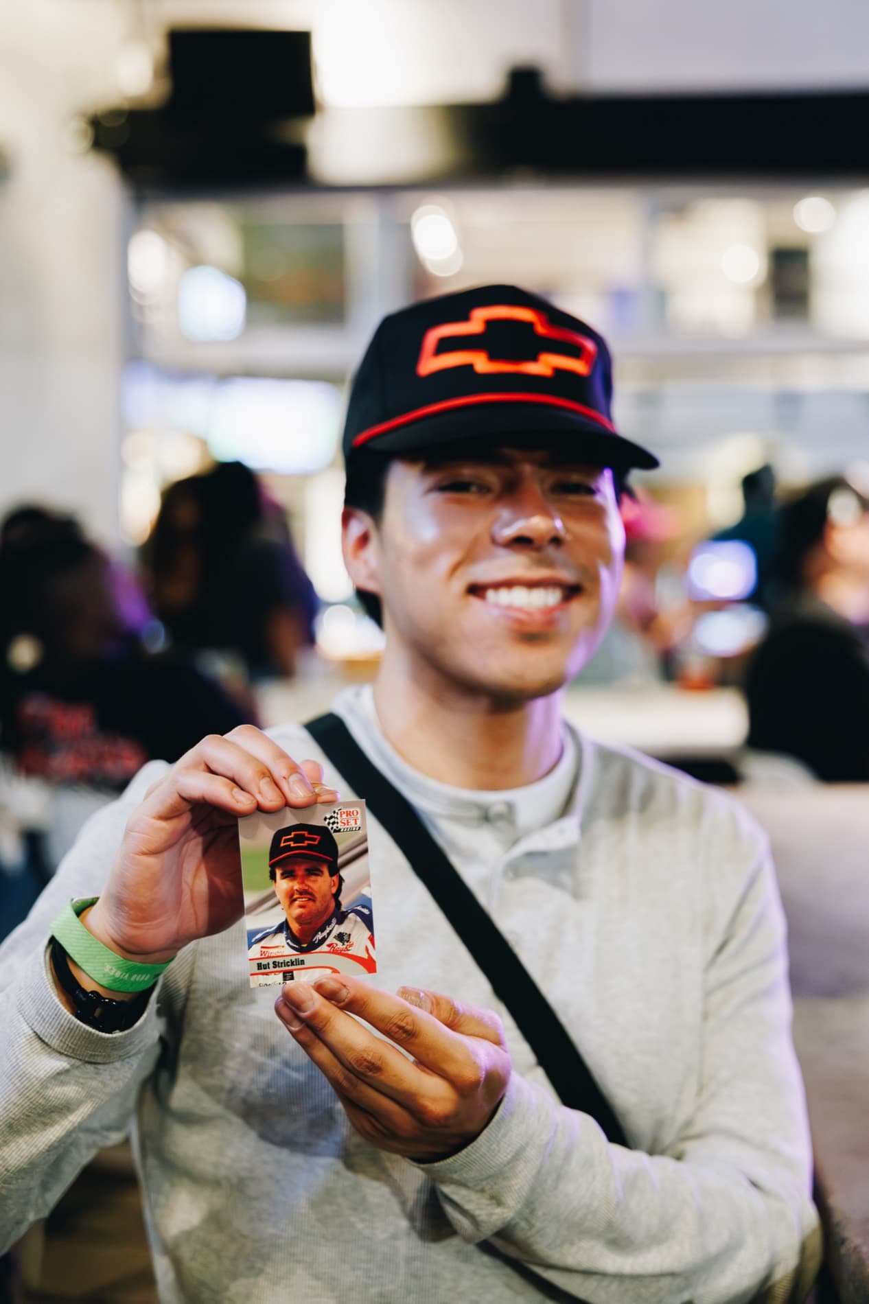 A smiling man wearing a black Chevrolet hat holds up a NASCAR trading card in a brightly lit indoor venue.