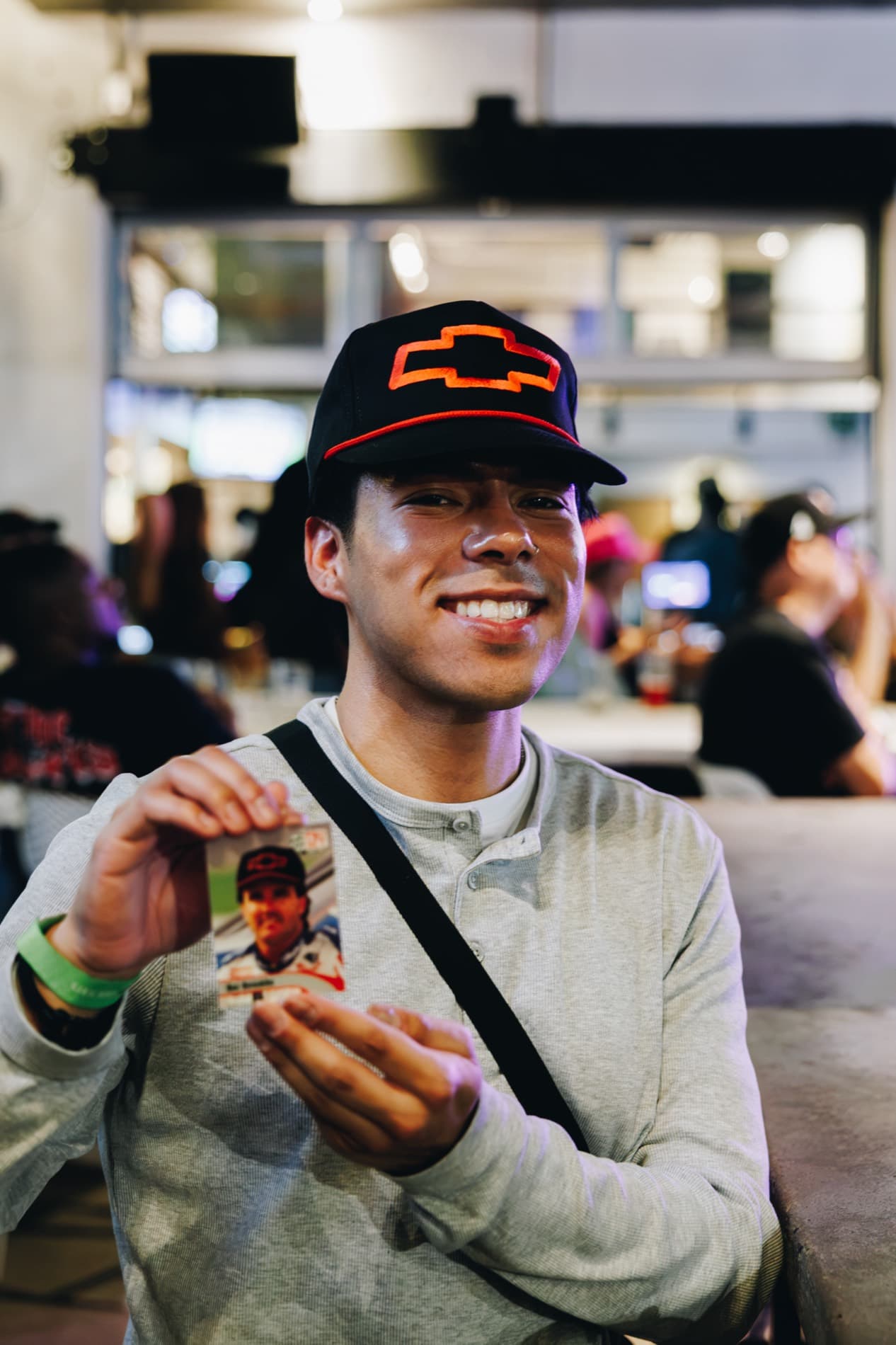 A smiling man wearing a black Chevrolet cap holds up a NASCAR trading card in a crowded indoor venue.