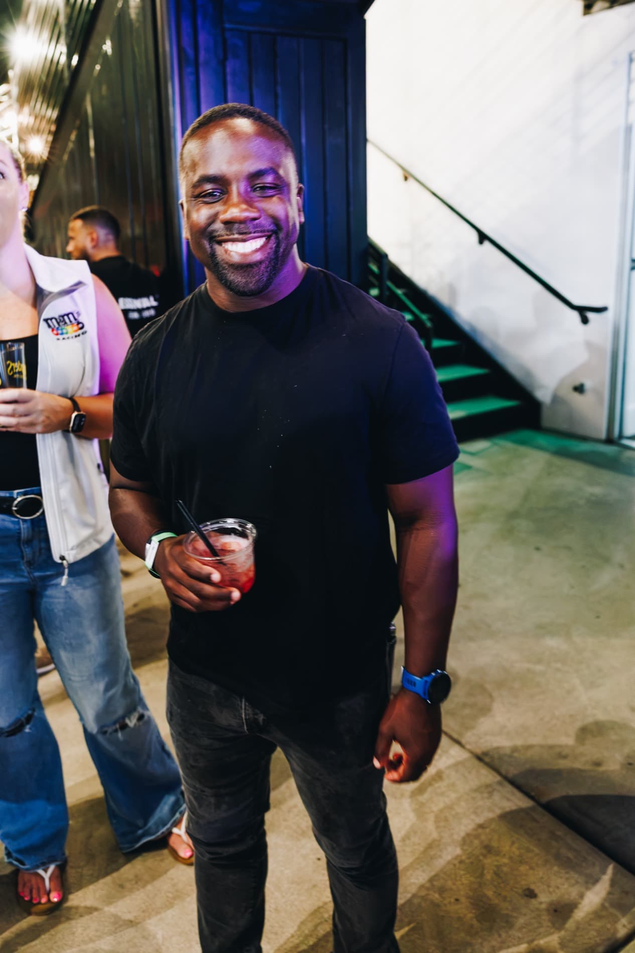 A smiling man in a black T-shirt holds a drink while standing in a lit indoor venue near stairs and other attendees.