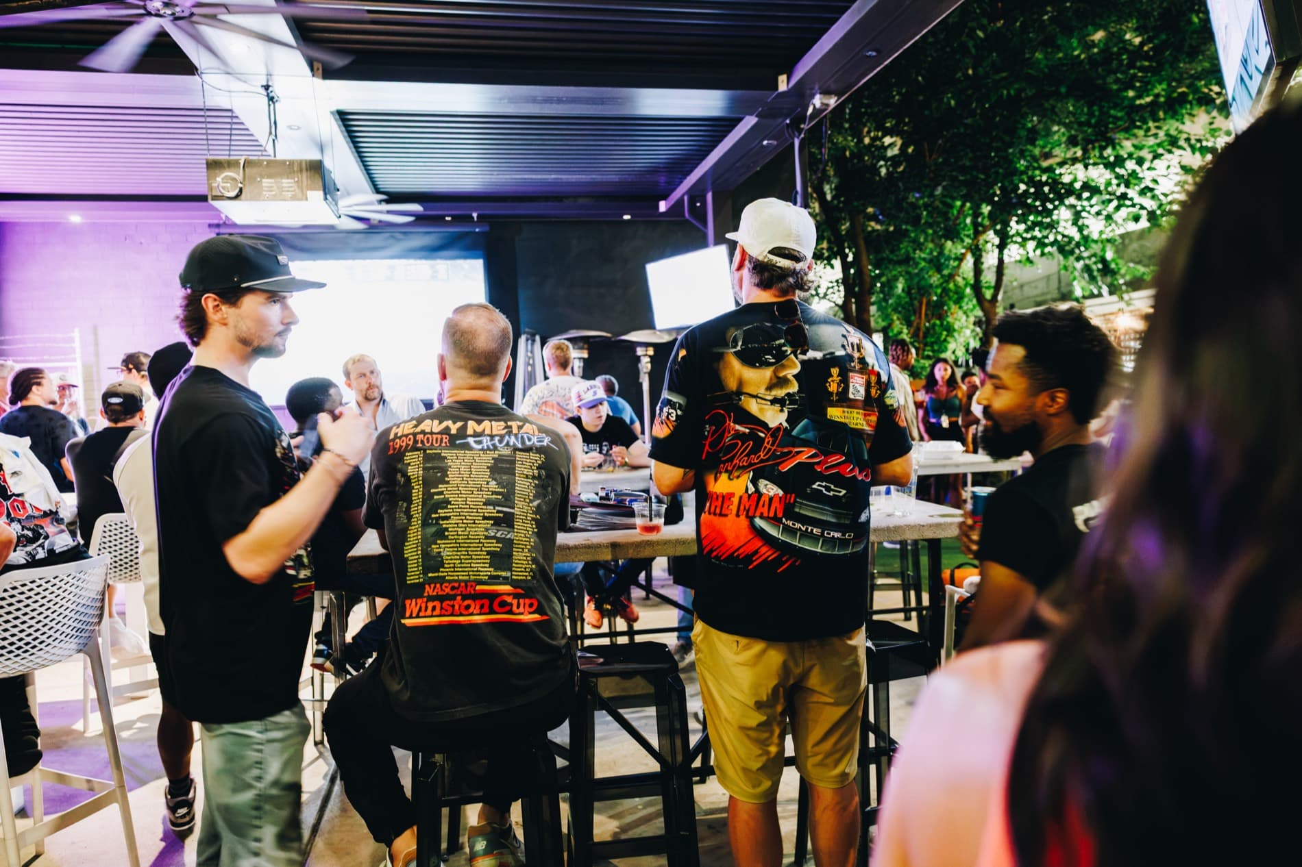 People in NASCAR-themed shirts gather at an outdoor bar or patio during a watch party.