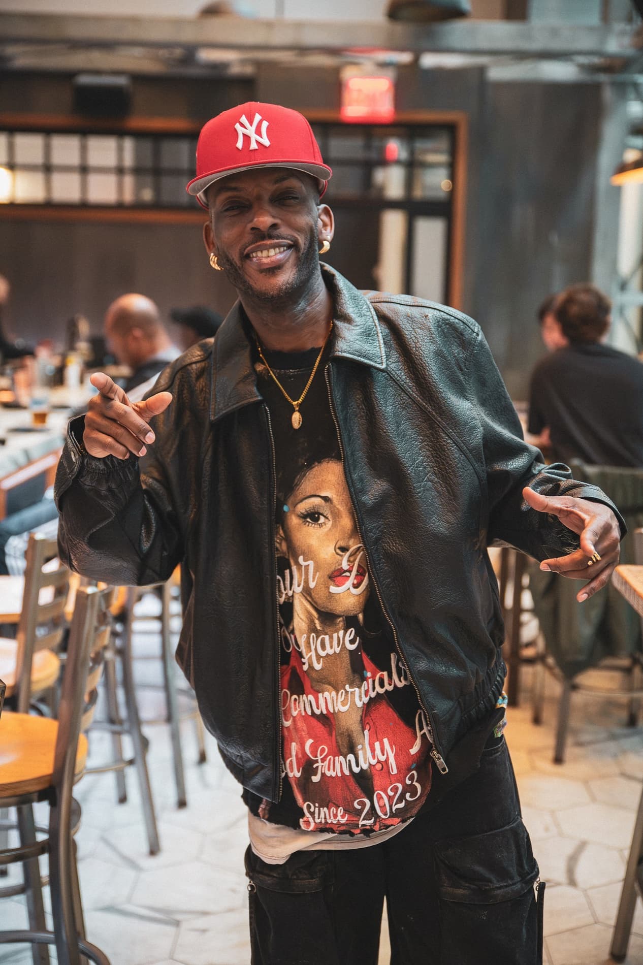 A smiling man in a red New York Yankees cap and a leather jacket points at the camera inside Burgerology.