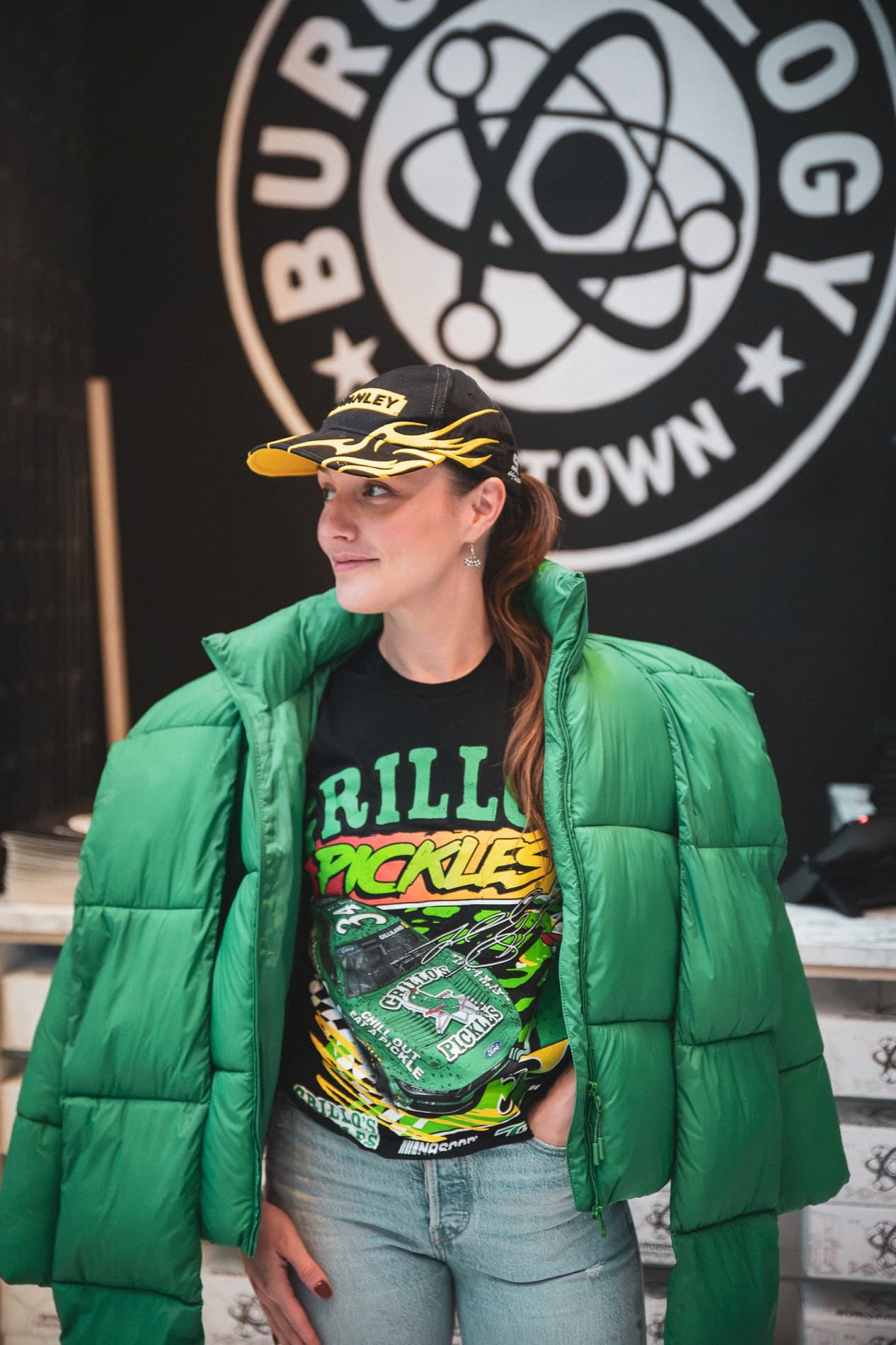 A woman in a green puffer jacket and a Grillo's Pickles race shirt stands in front of the Burgerology Midtown logo.