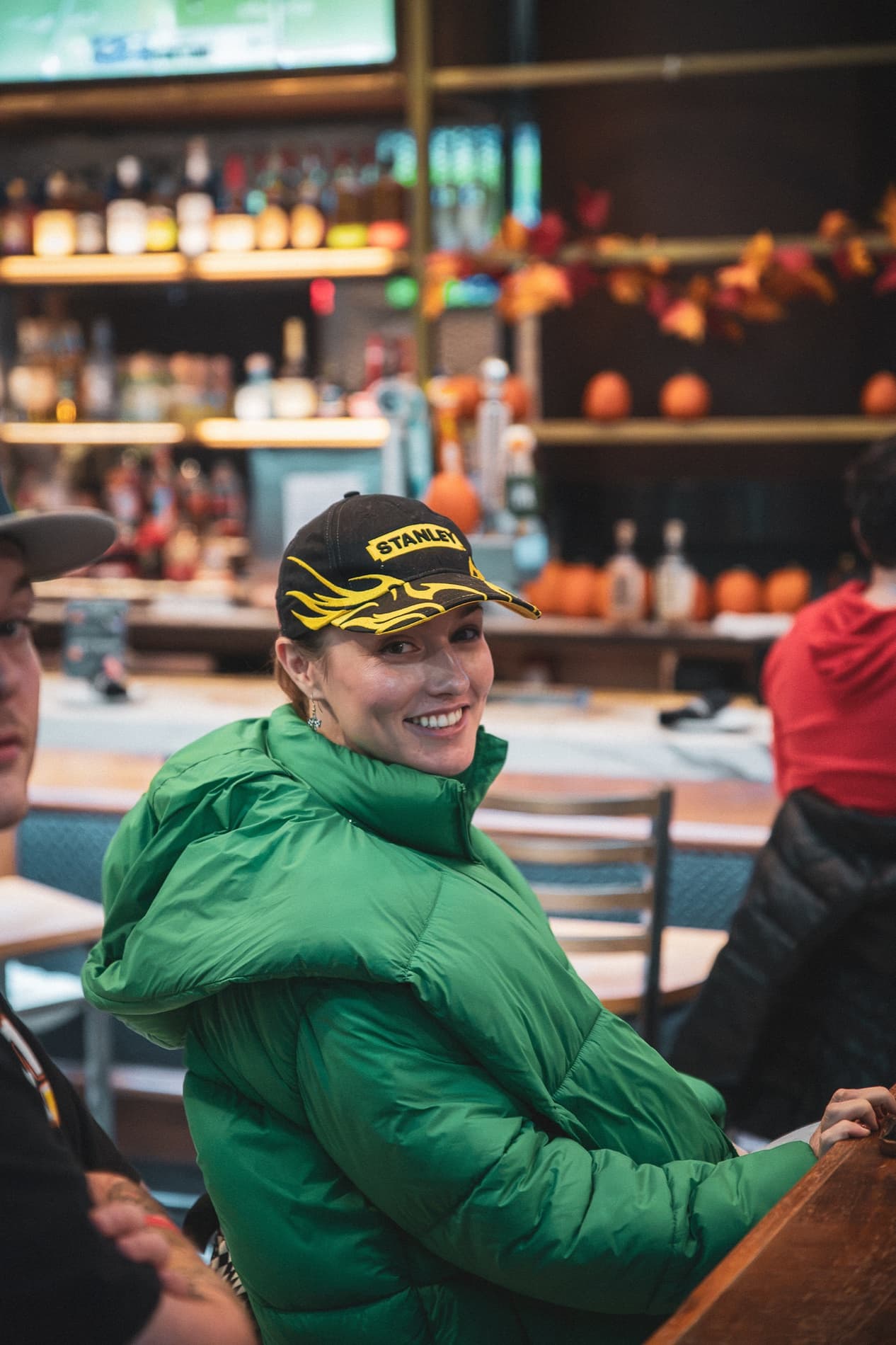 A smiling woman in a green puffer jacket and a race cap sits at the bar.