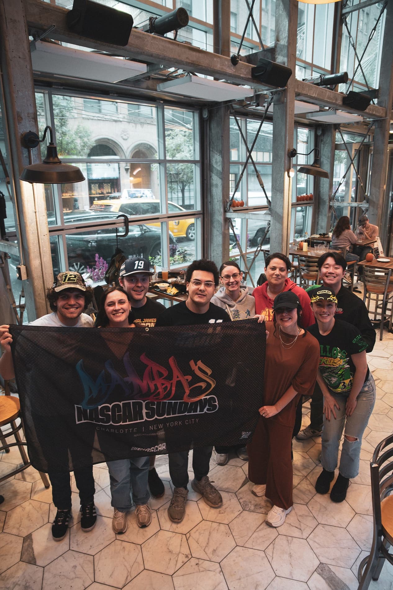 The group holds up the Mamba's NASCAR Sundays banner, seen from above, with the rainy street through the windows.