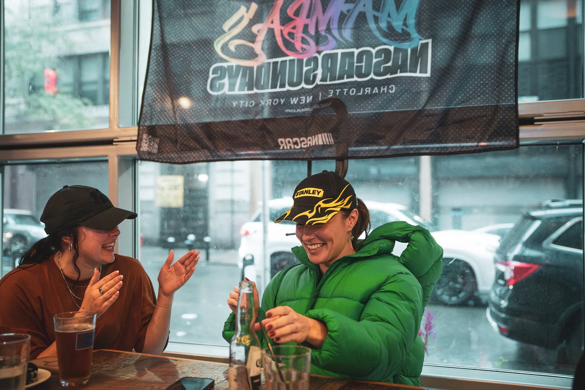 Two women laugh at a window table beneath the NASCAR Sundays banner as rain falls outside.
