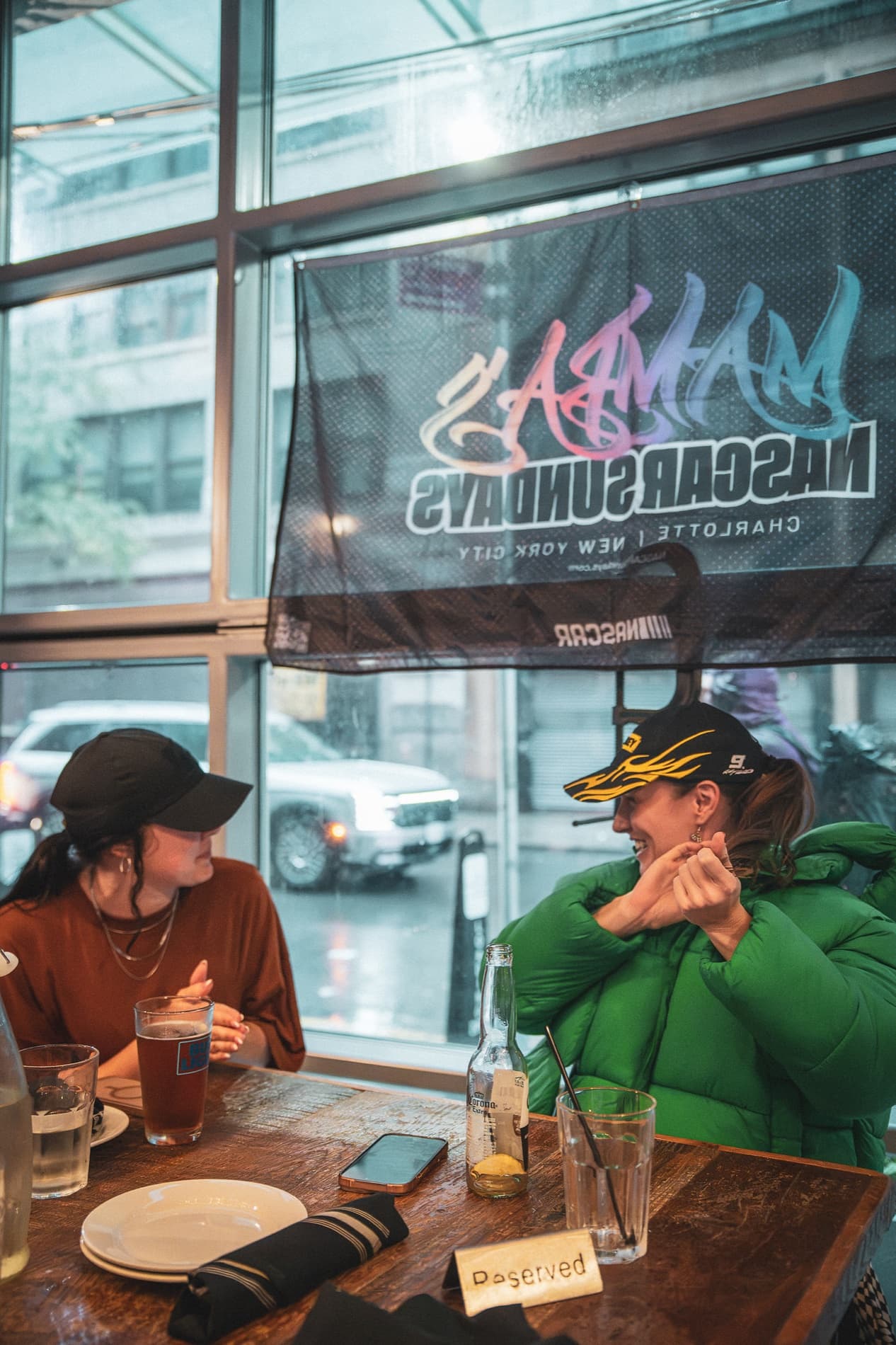 Two women at a window table under the NASCAR Sundays banner, one in a green jacket and race cap.
