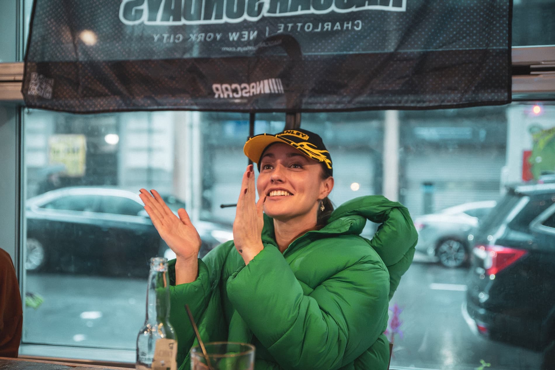 A woman in a green jacket and race cap claps beneath the NASCAR Sundays banner.