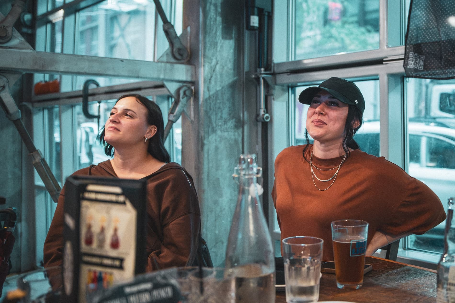Two women at a window table look up at the screens, one with a hand on her hip.