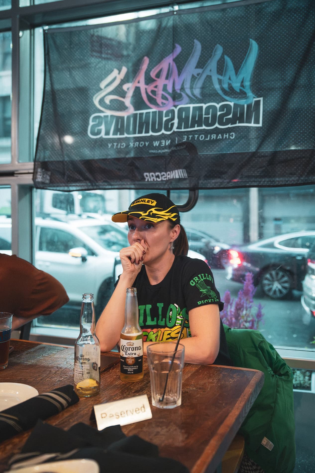 A woman in a race cap and Grillo's Pickles shirt watches with a hand at her mouth, the NASCAR Sundays banner in the window.